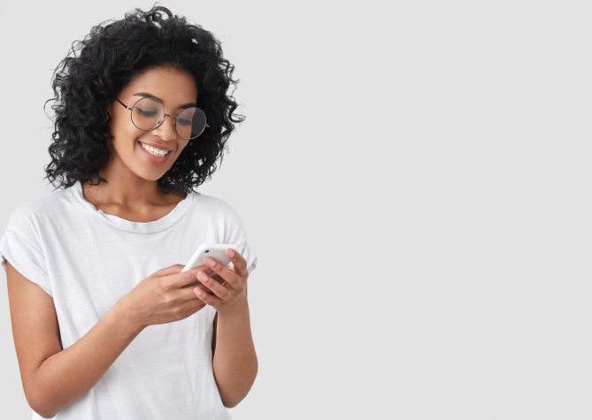 Cheerful African American with crisp hair, holds modern smart phone, happy to recieve message, dressed in casual t shirt, poses against white background with blank space for your advertisement