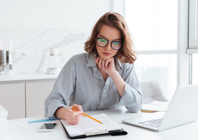 Young concentrated businesswoman in glasses and striped shirt working with papers at home