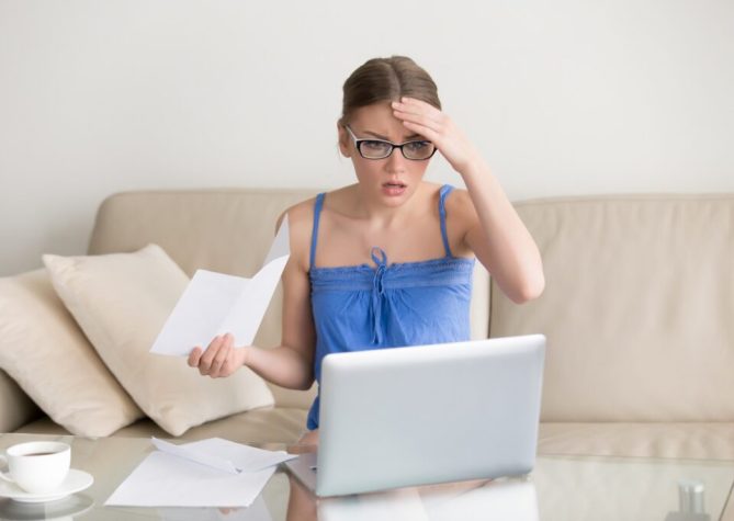 Worried young woman in eyeglasses sitting on sofa with paper letter in hand and looking with confusion on laptop screen. Female taxpayer with overdue tax returns shocked because of financial penalties