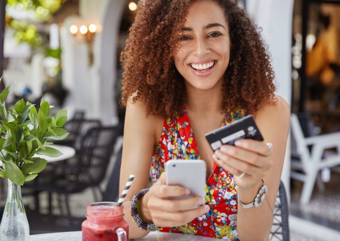 Internet banking and ecommerce concept. Happy young smiling female with Afro hairstyle, uses modern cell phone and credit card for online shopping, enjoys fresh fruit smoothie in terrace bar.