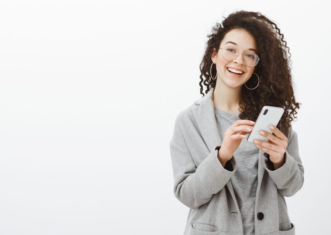 Joyful emotive young european woman in glasses and grey coat with stylish round earrings, holding smartphone and smiling cheerfully at camera while retelling interesting article to friend. Copy space