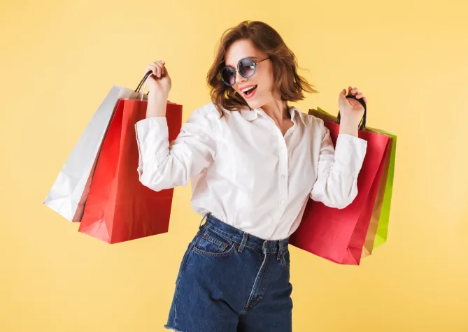 Portrait of happy lady in sunglasses standing with colorful shopping bags in hands on over pink background. Young woman standing in white shirt and denim shorts